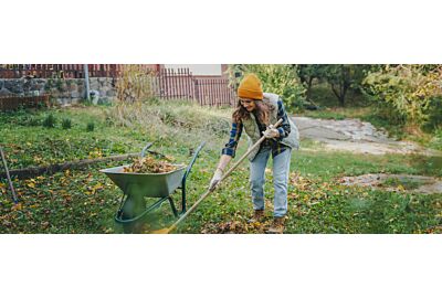 kobieta ubrana w kamizelkę i jesienne spodnie grabiąca liście w ogrodzie