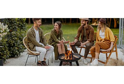 Kobieta z psem spędzająca czas w ogrodzie przy palenisku ogrodowym ze szklarnią w tle, obok wygodny biały fotel z technorattanu