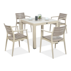 Beżowy stół kwadratowy z kompletem czterech krzeseł w tym samym kolorze, ustawionych wokół, na blacie dekoracja w szklanym kloszu i filiżanki.