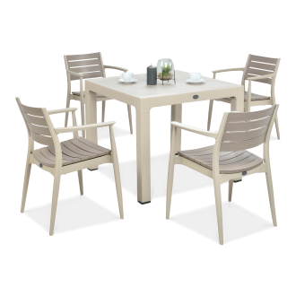 Beżowy stół kwadratowy z kompletem czterech krzeseł w tym samym kolorze, ustawionych wokół, na blacie dekoracja w szklanym kloszu i filiżanki.