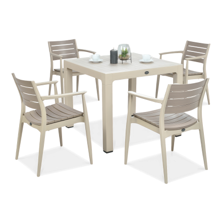 Beżowy stół kwadratowy z kompletem czterech krzeseł w tym samym kolorze, ustawionych wokół, na blacie dekoracja w szklanym kloszu i filiżanki.