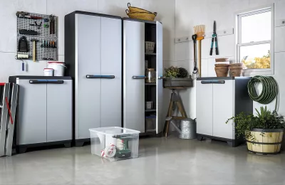 Zestaw szaf ogrodowych w jasnoszarej kolorystyce ustawionych w warsztacie, na ścianie narzędzia i akcesoria ogrodnicze.