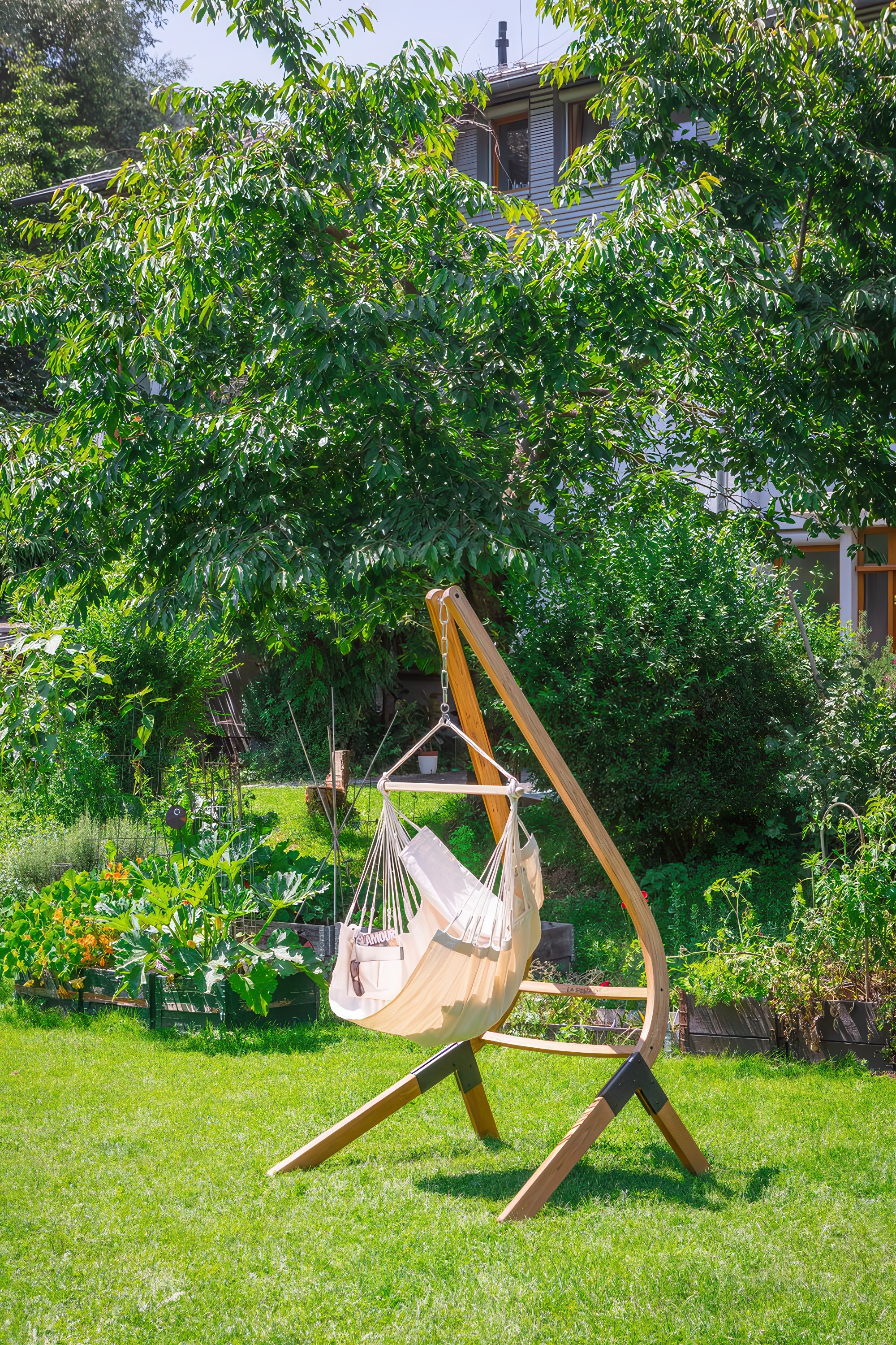 Fotel hamakowy HABANA Basic Latte z bawełny organicznej, zawieszony na drewnianym stojaku, umieszczony w ogrodzie na tle zielonych drzew i krzewów.

