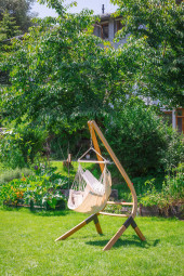 Fotel hamakowy HABANA Basic Latte z bawełny organicznej, zawieszony na drewnianym stojaku, umieszczony w ogrodzie na tle zielonych drzew i krzewów.
