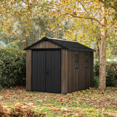 Domek na narzędzia Keter Deco Apex 7511/1175 w kolorze orzecha z czarnymi drzwiami i oknami otoczony jesienną roślinnością