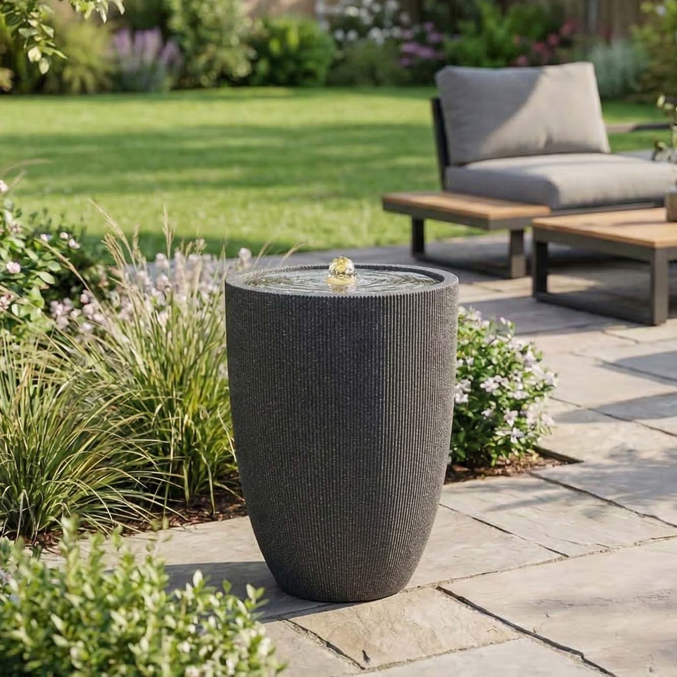 Fontanna ogrodowa grafitowa ustawiona na tarasie w otoczeniu roślin i mebli ogrodowych
