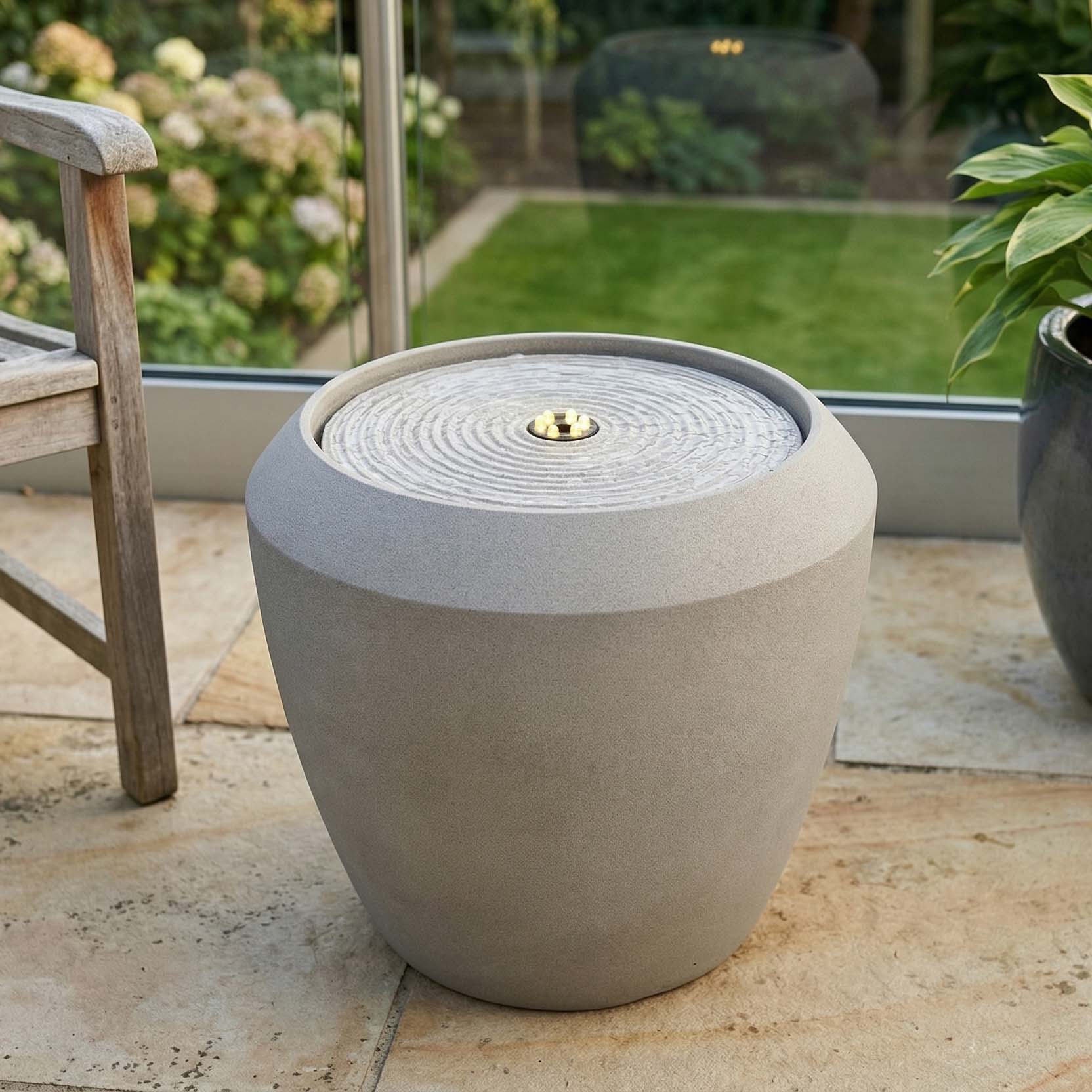 Nowoczesna fontanna ogrodowa w aranżacji na tarasie obok roślin i drewnianego krzesła
