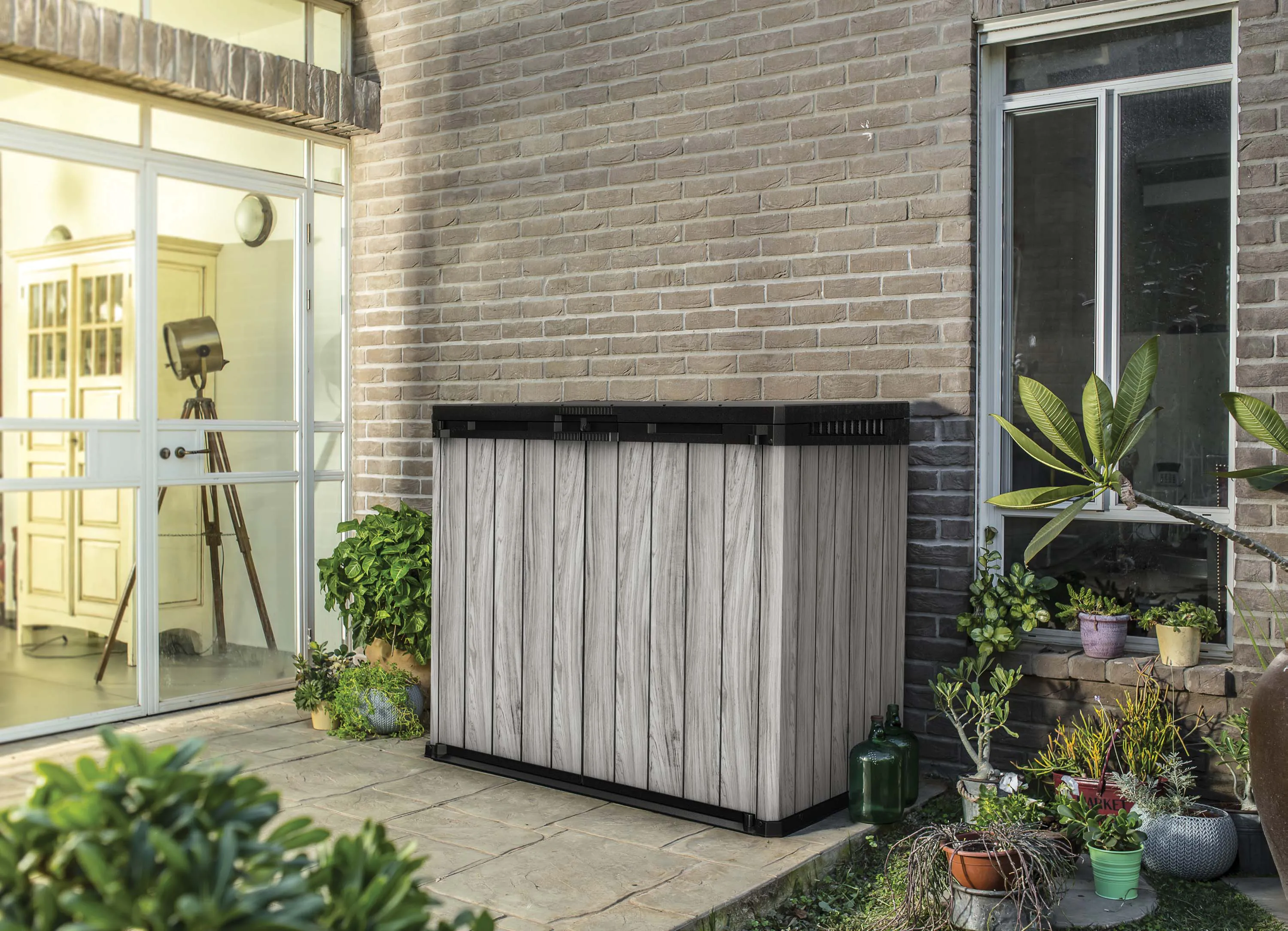 Szary organizer ogrodowy SIO Darwin 1150L Storm Grey marki Keter postawiony przy ceglanej ścianie domu w ogrodzie