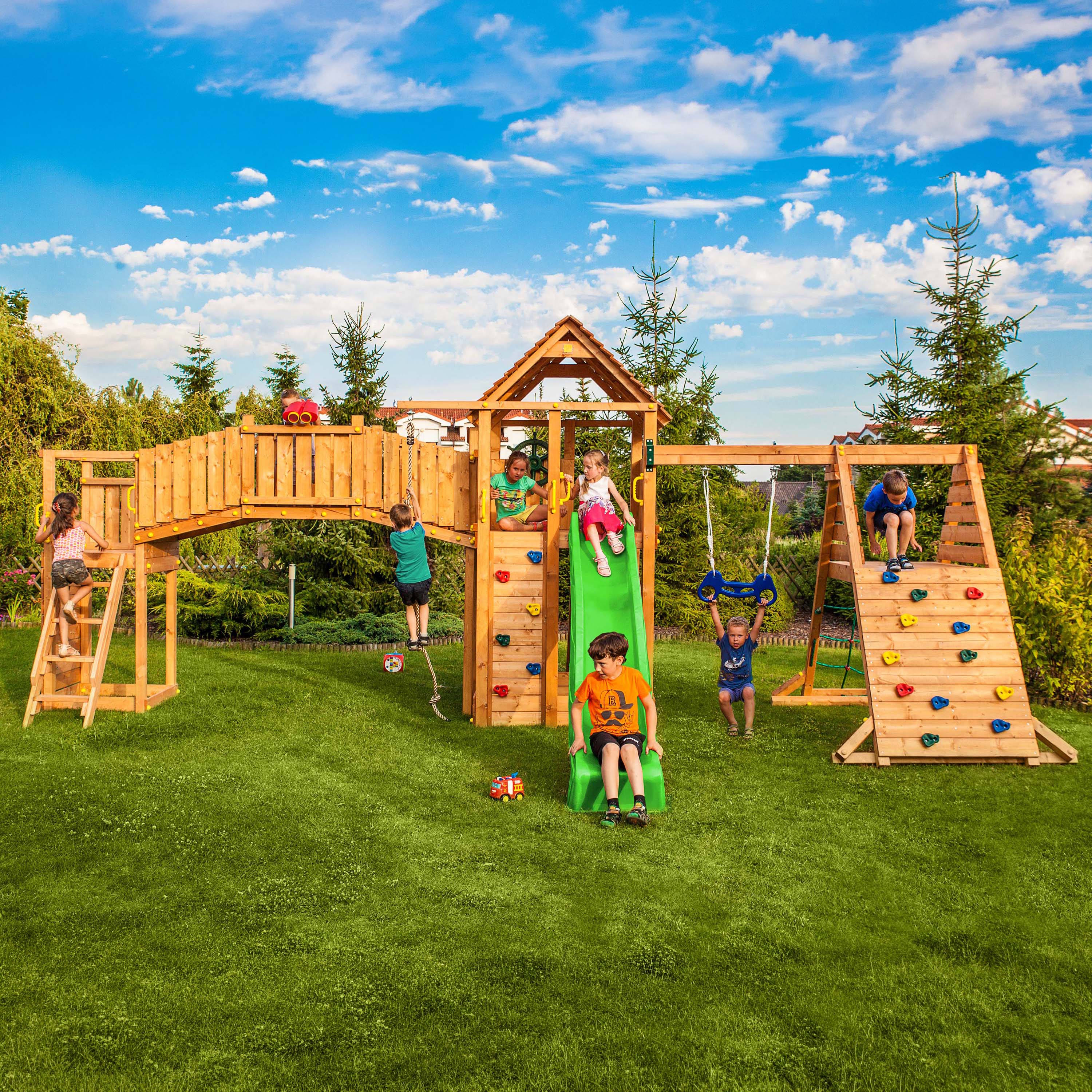 Duży drewniany plac zabaw w ogrodzie z mostkiem zjeżdżalnią huśtawką i ścianką wspinaczkową
