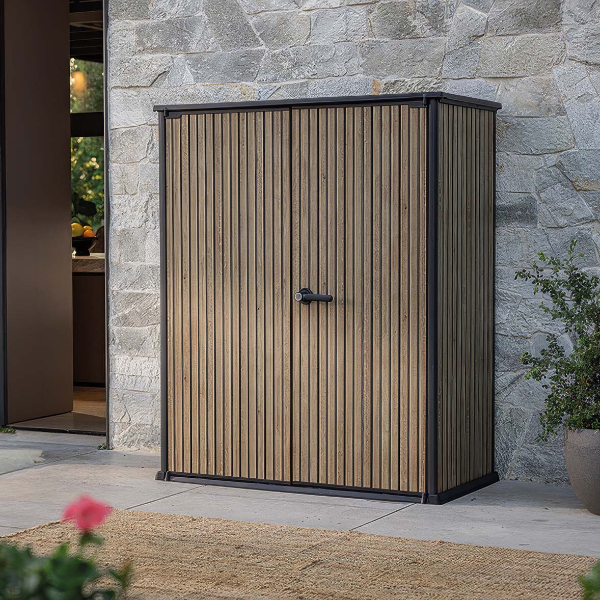 Schowek ogrodowy Deco Vertical 1400L Venetian Brown Keter z dwuskrzydłowymi drzwiami na tle kamiennej ściany budynku