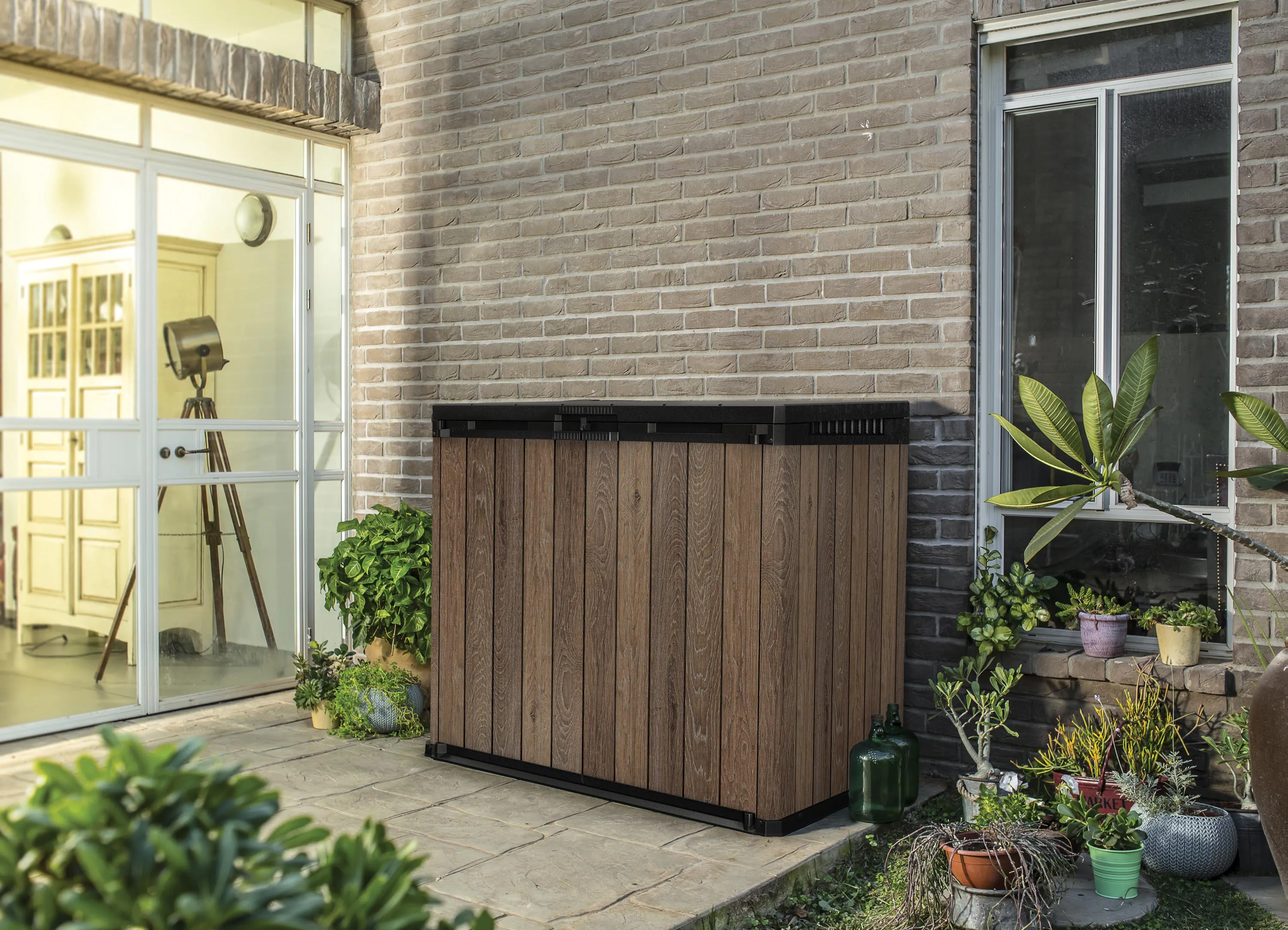 Szafa ogrodowa SIO Darwin 1150l Walnut w kolorze brązowym ustawiona na tarasie przy ceglanej ścianie domu