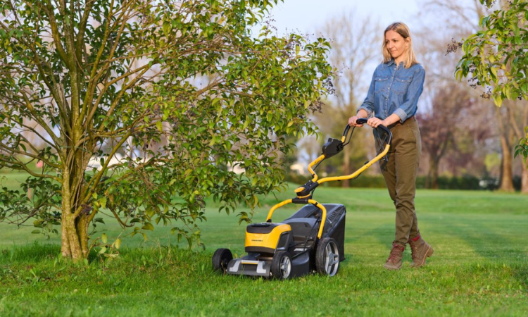 kobieta kosząca trawę pod drzewami ż&oacute;łtą kosiarką 