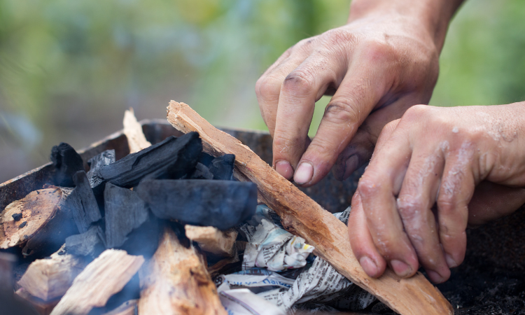 dłonie meżczyzny rozpalajace grilla z brykietem, gałązkami i papierem