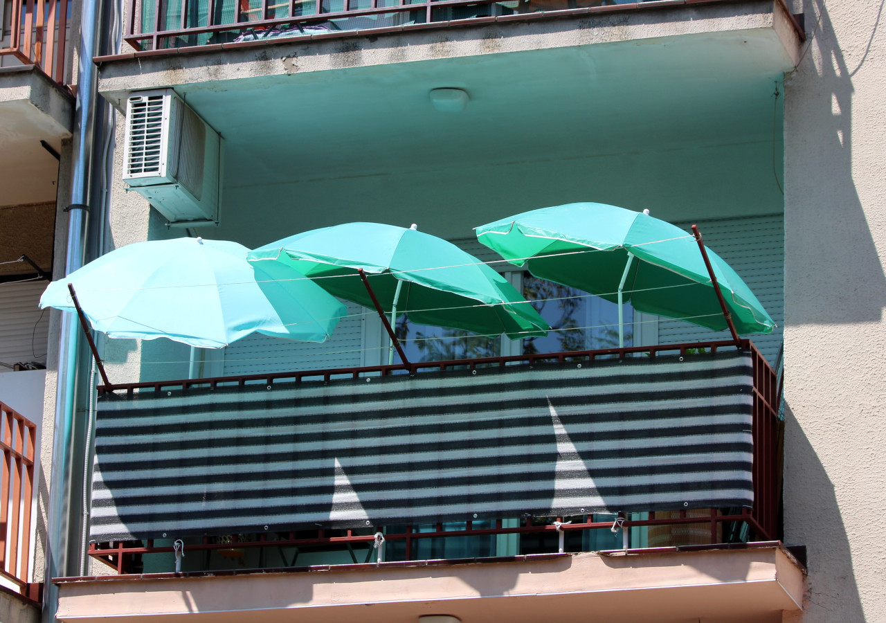 Balkon w bloku osłonięty matą balkonową w pasy, z trzema zielonymi parasolami przeciwsłonecznymi