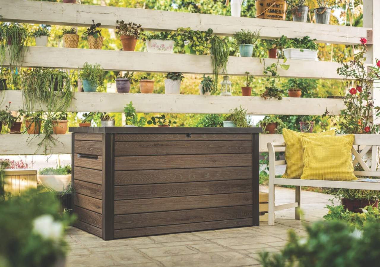 Drewniana skrzynia ogrodowa ustawiona na tarasie, obok białej ławki z żółtymi poduszkami, w otoczeniu donic z roślinami i kwiatami.