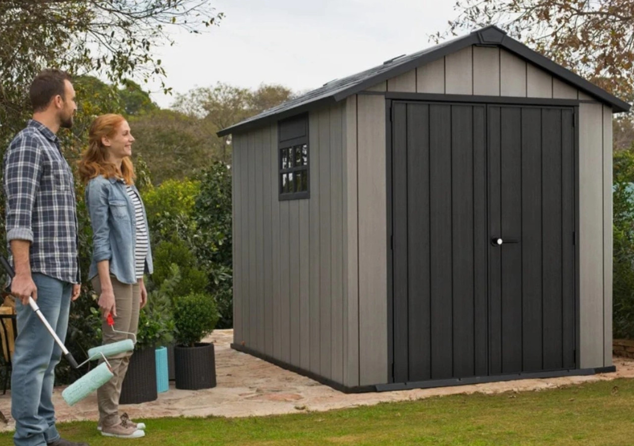 Domek narzędziowy OAKLAND 759 - 279x210cm - KETER w ogordzie, obok domku stoi kobieta i mężczyzna z narzędziami ogrodowymi 
