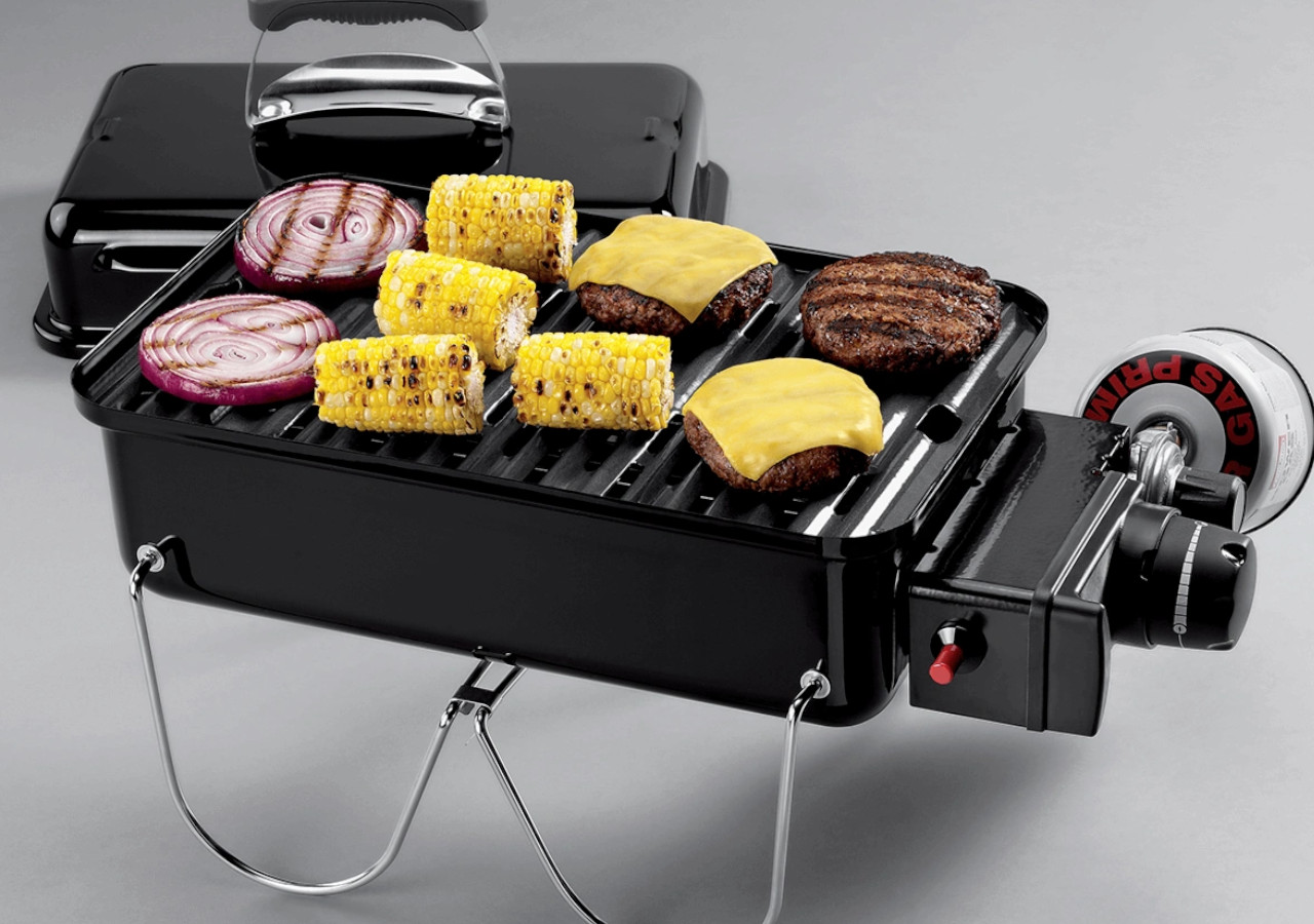 Grill gazowy Go-Anywhere - WEBER na szarym tle, w środku kukurydza i inne jedzenie przygotowane na grilla