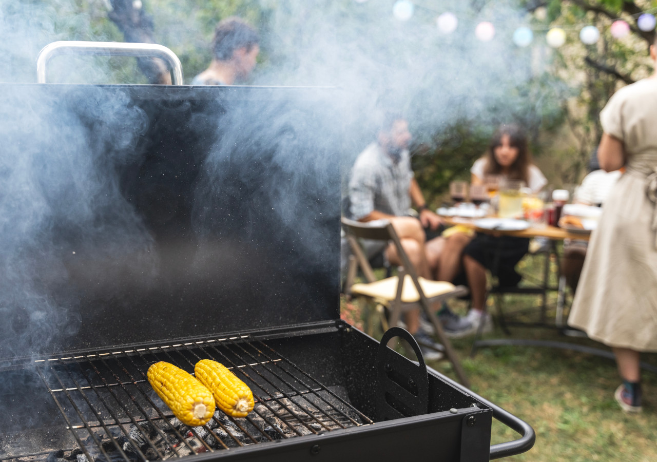 Kukurydza grillowana na ruszcie z dymem unoszącym się w powietrzu, w tle grupa osób siedzących przy stole w ogrodzie podczas letniego spotkania.