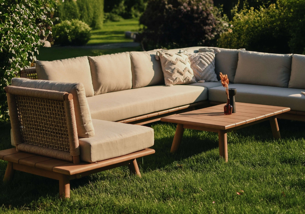 Beżowy narożnik ogrodowy z poduszkami i drewnianym stolikiem na trawie w słonecznym ogrodzie.