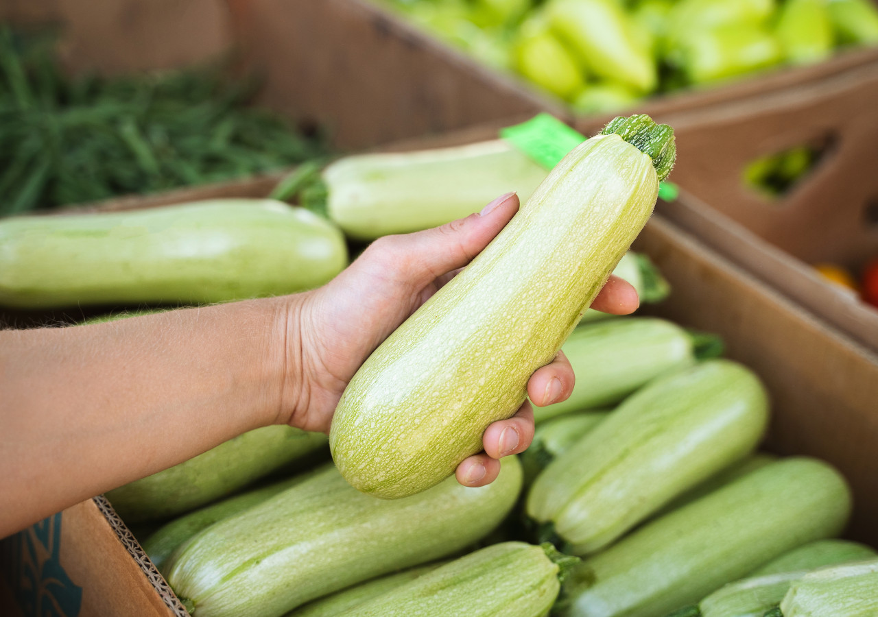Osoba trzyma w dłoni świeżą cukinię nad skrzynką pełną warzyw na targu