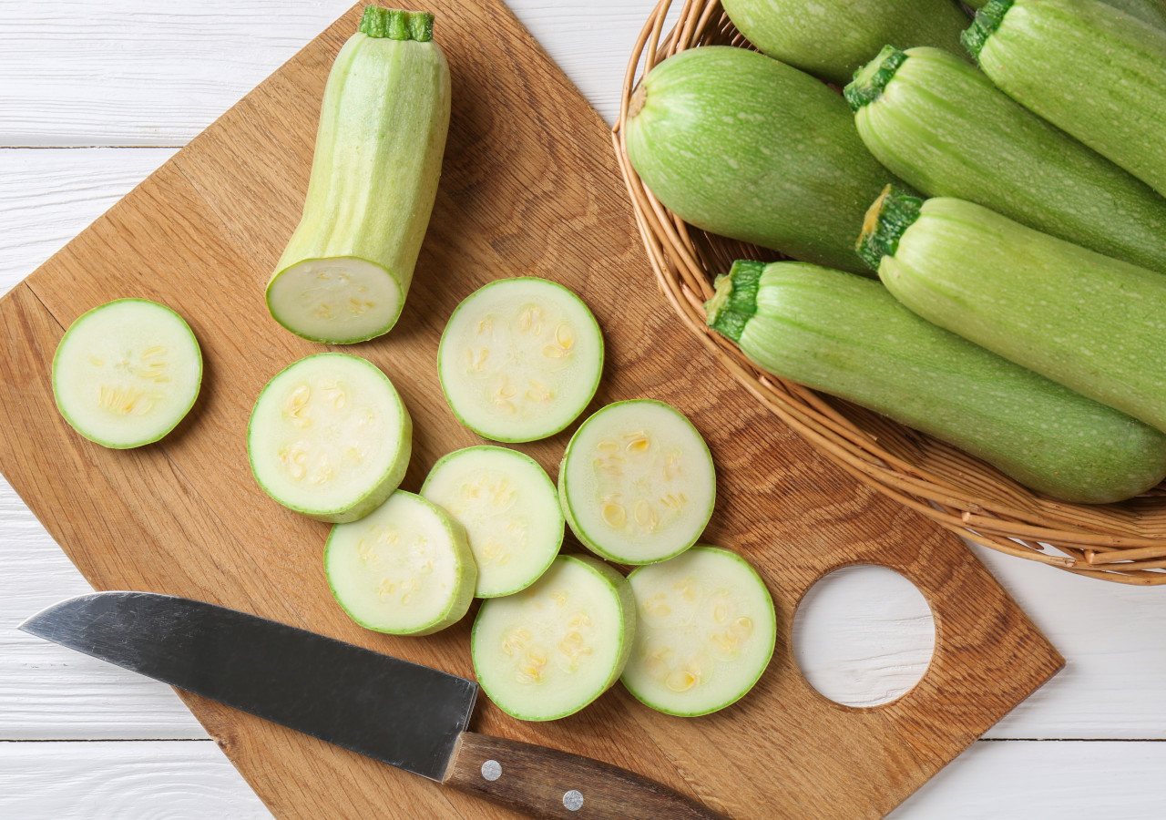 Pokrojona cukinia na drewnianej desce do krojenia z nożem, obok koszyka pełnego świeżych cukinii.