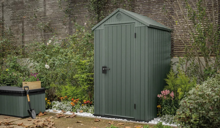 Domek ogrodowy DARWIN 4x4 zielony - Keter w ogrodzie pod płotem okalany zielenią i kwiatami