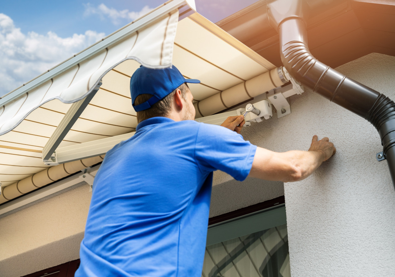 Pracownik w niebieskim stroju montuje markizę tarasową na elewacji domu.